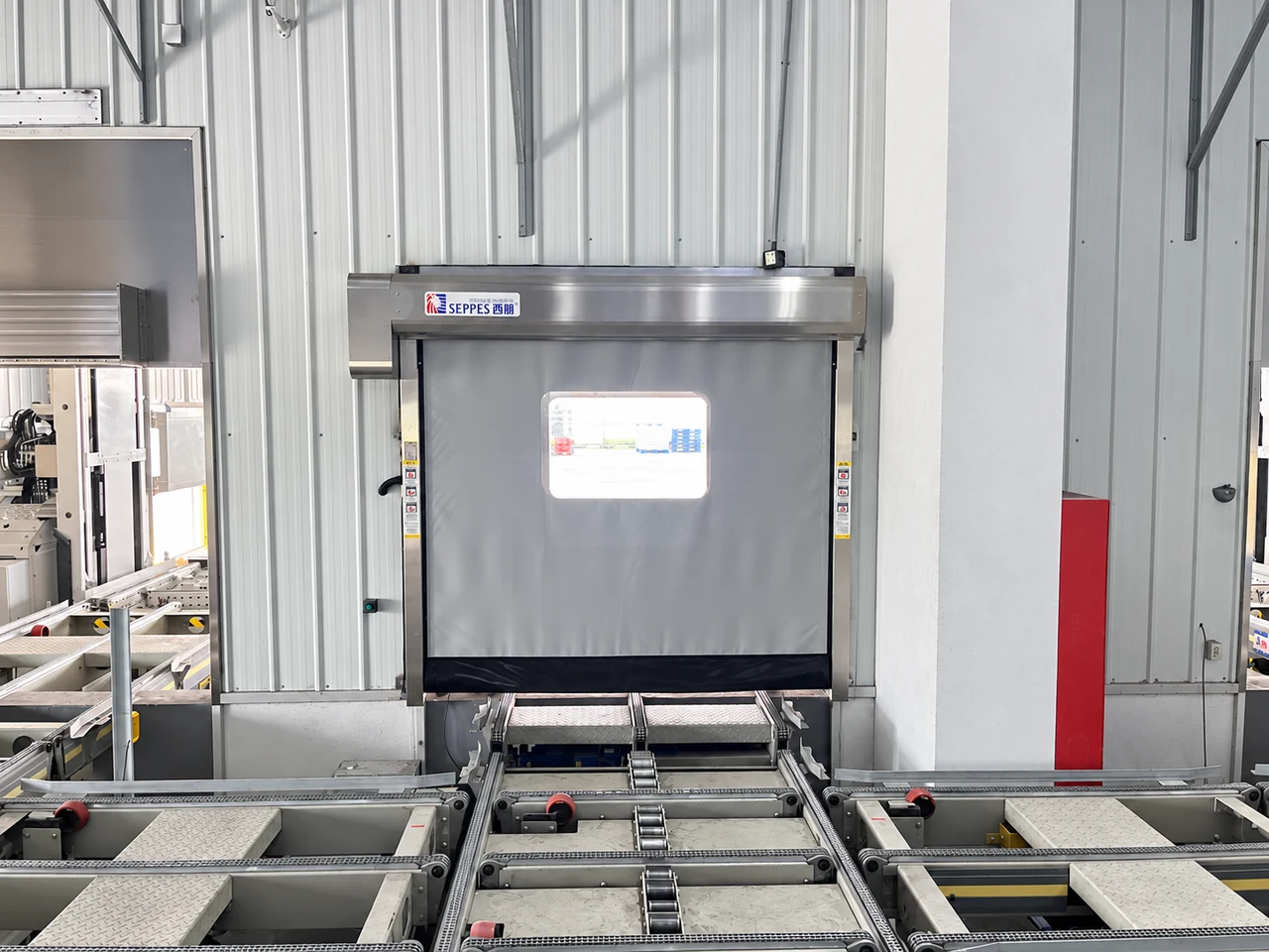Grey SEPPES fabric roll-up door mounted over a conveyor transfer opening in a production facility