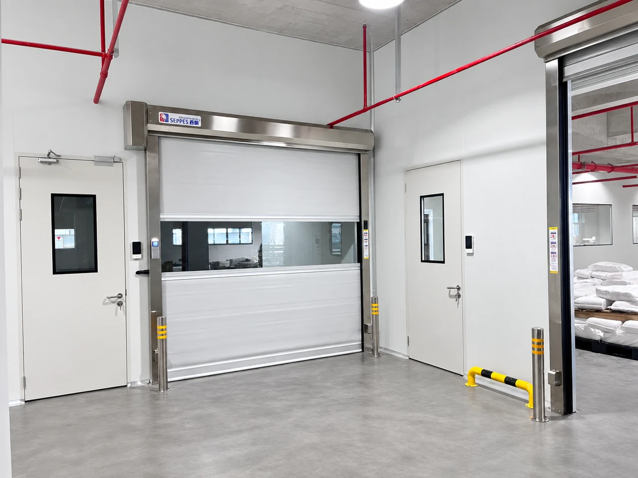 White brush-sealed high speed fabric door beside personnel doors, with a vision panel and protective bollards